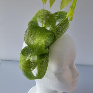 May include: A lime green headpiece with a woven texture and upright feathers. The headpiece is mounted on a white mannequin head. The feathers are a lighter shade of green. The headpiece is a formal accessory.