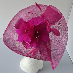 Pode incluir: Um fascinator rosa fúcsia com uma flor grande e penas. A flor tem algumas pequenas pedras de cristal no centro.