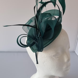 Puede incluir: Un fascinador verde oscuro con plumas y red, sujeto a una diadema.