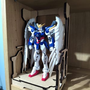 Può includere: Una figura modello Wing Gundam con armatura bianca, blu e rossa e grandi ali piumate bianche, in piedi su una base espositiva in legno tagliato al laser con una struttura a telaio abbinata che sostiene la figura in posizione eretta.