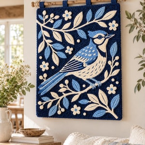 Puede incluir: Un tapiz rectangular de punch needle que muestra un arrendajo azul posado en una rama. El diseño incluye hojas y flores en color crema y azul claro sobre un fondo azul marino intenso. Está suspendido de una varilla de madera con una cuerda.