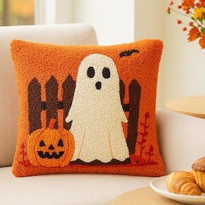 Puede incluir: Cojín decorativo naranja con temática de Halloween con un fantasma blanco, una calabaza y una valla marrón. El cojín está colocado en un sofá beige. Un jarrón con flores naranjas y una mesa con pasteles están en el fondo.