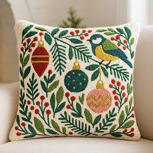 May include: Square cushion with a cream background featuring embroidered Christmas decorations, a bird, and green foliage. The decorations are red, blue, and pink. The bird has yellow, blue, and green feathers. The cushion is on a white chair.