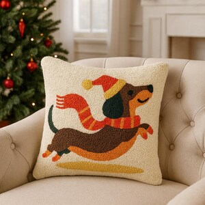 Peut inclure: Un coussin carré de couleur crème avec un motif en punch needle texturé représentant un teckel marron. Le chien porte un bonnet d'hiver et une écharpe rayés rouge et jaune. Le motif est réalisé avec une texture de laine bouclée en relief.