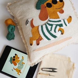 May include: A square beige throw pillow with a fringed edge featuring a punch needle design of a brown dachshund wearing a green beret, black sunglasses, and a white and green striped sweater. Includes yarn, a tablet, fabric, and a needle tool.