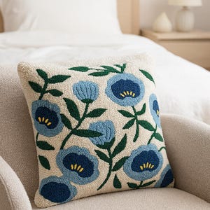 May include: A square decorative pillow with a cream-colored background and a floral pattern. The flowers are blue with yellow centers and green leaves. The pillow is placed on a beige chair.