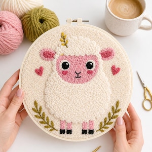Op de afbeelding: Een ronde schapenborduurring met een pluizig wit lichaam, roze gezicht, oren en harten. Het schaap heeft zwarte ogen en voeten en is omgeven door groene bladeren. Garenballen en een kop koffie staan op de achtergrond.