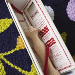 Może przedstawiać: Drewniane narzędzie do dziurkowania z czerwoną przędzą, w białym pudełku. Pudełko zawiera kartę "STITCH GAUGE" z instrukcjami do robienia dywanów. Tło to kolorowy, teksturowany materiał.