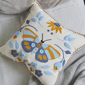 Puede incluir: Una almohada blanca con una mariposa bordada en azul y amarillo, flores y hojas. La almohada tiene un borde cosido decorativo.