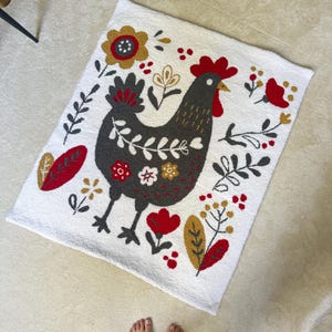 Könnte beinhalten: Ein weißer Teppich mit einem grauen und roten Hahn-Design. Der Hahn ist von stilisierten Blumen und Blättern in Rot-, Gelb- und Grüntönen umgeben.