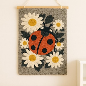 Könnte beinhalten: Ein gerahmtes Wandbild mit einem Marienkäfer- und Gänseblümchen-Design. Der Marienkäfer ist rot mit schwarzen Punkten, umgeben von weißen Gänseblümchen mit gelben Zentren und dunkelgrünen Blättern, alles vor einem grauen, strukturierten Hintergrund. Aufgehängt an einer Holzstange und einer Naturkordel.