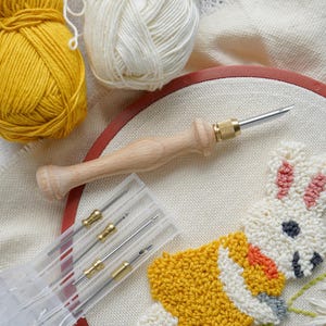 May include: A wooden punch needle tool with a metal needle and gold-colored accents rests on an embroidery hoop with a partially completed bunny design. Two balls of yarn, one yellow and one cream, are in the background, along with a clear plastic case containing additional needles.