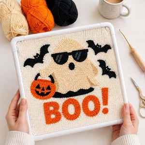 May include: A square punch needle embroidery project in a white frame featuring a beige ghost wearing black sunglasses, holding an orange jack-o'-lantern bucket, surrounded by black bats, with the orange text BOO! at the bottom.