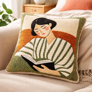 May include: A decorative cushion with an illustration of a person reading. The cushion has a cream background, green border, and a striped shirt design. The person's face is peach-coloured with black hair. The cushion is on a beige sofa.