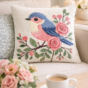 May include: A decorative cushion with a textured white background. The cushion features an embroidered design of a blue and pink bird on a branch with pink roses and green leaves. The cushion is on a beige sofa.