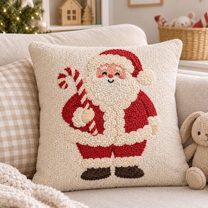 Könnte beinhalten: Ein quadratisches Zierkissen mit cremefarbener, strukturierter Oberfläche und einem Punch-Needle-Design des Weihnachtsmanns. Er trägt einen roten Anzug mit cremefarbenem Besatz, hält eine rot-weiß gestreifte Zuckerstange und trägt dunkelbraune Stiefel.