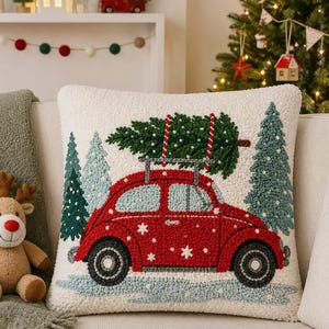 Puede incluir: Un cojín cuadrado con fondo color crema que presenta un diseño texturizado de aguja mágica de un coche rojo antiguo que lleva un árbol de Navidad verde en el techo, rodeado de pinos verdes y copos de nieve blancos sobre un suelo azul claro.