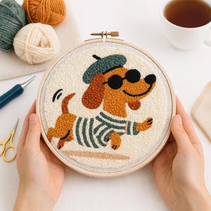 Puede incluir: Un bastidor de bordado con aguja perforadora con un perro salchicha de dibujos animados que lleva gafas de sol, una boina y una camisa a rayas. El perro es marrón y canela con detalles en negro. El fondo es de tela color crema. El bastidor se sostiene en las manos.