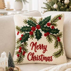 May include: A square throw pillow with a cream-coloured textured base featuring a punch needle design of green holly leaves, pine branches, and red berries. The text Merry Christmas is embroidered in red script lettering across the centre.