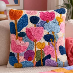 May include: A square throw pillow featuring a textured punch needle design of abstract flowers in vibrant shades of orange, pink, blue, yellow, purple, and green against an off-white background. The surface has a raised, looped yarn texture.
