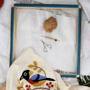 May include: An embroidery project in progress, featuring a wooden frame with a stretched white fabric. A colorful bird design is partially stitched on the fabric. Additional supplies include a ball of yarn, scissors, and a punch needle tool.
