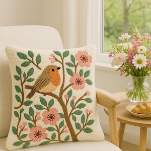 Peut inclure: Coussin carré crème brodé d'un oiseau perché sur une branche d'arbre avec des fleurs roses et des feuilles vertes. Le coussin est posé sur une chaise crème, près d'une fenêtre.