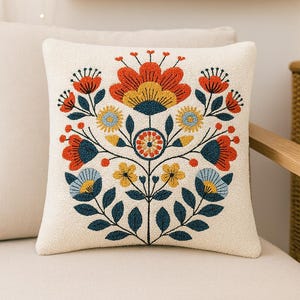 May include: Cream-coloured square cushion with a colourful floral design. The embroidered pattern features a large orange flower at the top, surrounded by blue, yellow, and red flowers and green leaves. The cushion is on a cream-coloured chair.