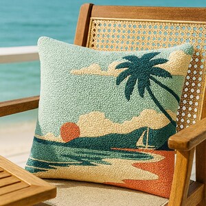 May include: Square decorative pillow with a beach scene design. The pillow features a palm tree, sailboat, and sunset in shades of green, orange, and blue. The pillow is placed on a wooden chair with a woven back, with the ocean in the background.