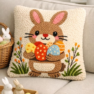 May include: A decorative Easter-themed pillow featuring a brown bunny holding colorful Easter eggs. The pillow has a cream-colored background with a textured, tufted design. The bunny has pink ears and a red nose. Decorative plants are on either side.