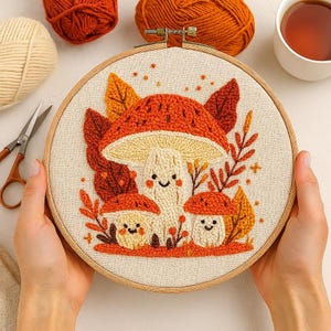 May include: A mushroom-themed embroidery hoop art piece. The design features three smiling mushrooms in shades of orange, cream, and beige, surrounded by fall leaves. The artwork is set in a wooden hoop, with yarn and scissors in the background.