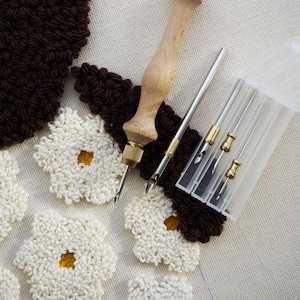Puede incluir: Primer plano de un proyecto de artesanía en curso, con una herramienta de aguja de perforación de madera, agujas y flores de hilo blanco y marrón sobre un lienzo. Las flores tienen centros amarillos.