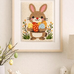 May include: Framed Easter-themed artwork featuring a brown bunny holding colorful Easter eggs. The artwork is in a light wooden frame and has a textured, stitched appearance. The bunny is surrounded by green and yellow floral accents, creating a festive spring scene.