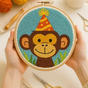 Può includere: Opera d'arte ricamata a mano su telaio raffigurante una scimmia sorridente con un cappello da festa. La scimmia è marrone, beige e gialla, su sfondo blu. L'opera è incorniciata in un telaio di legno, con erba verde in basso.