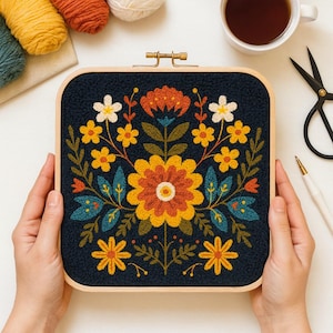 May include: A square embroidery hoop featuring a floral design in yellow, orange, and white on a navy blue background. The wooden frame is held by hands, with yarn, scissors, and a cup of tea visible.