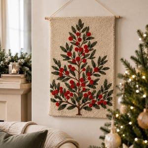 May include: A rectangular wall hanging featuring a punch needle design of a tree with dark green leaves and bright red berries on a cream-coloured background. The textile art is suspended from a wooden dowel with a thin string for wall mounting.