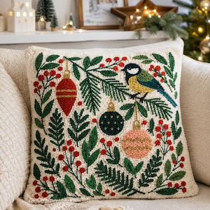 May include: A square throw cushion with a cream-coloured base featuring a textured punch needle design. The pattern displays a bird perched on pine branches, surrounded by red berries, a red ornament, a dark blue ornament, and a pink ornament.