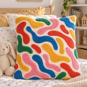 Puede incluir: Un cojín cuadrado con un diseño texturizado de aguja mágica que presenta un patrón abstracto de formas orgánicas onduladas en tonos vibrantes de rojo, azul, amarillo, verde y rosa sobre una tela de fondo color crema.