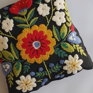 Peut inclure: Un coussin carré noir orné d'une broderie à l'aiguille perforée représentant diverses fleurs aux tons rouge, jaune, blanc et bleu, accompagnées de feuilles et de tiges vertes, formant un motif floral dense et texturé sur toute la surface.