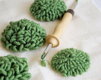 May include: Green yarn pom-poms on a white canvas with a wooden yarn punch tool.