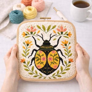May include: Square embroidery hoop featuring a detailed beetle design with floral accents. The beetle has a black body and yellow wings with pink floral patterns. The background is a textured cream color, surrounded by a wooden frame.