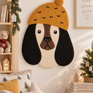 Peut inclure: Une tapisserie murale en punch needle représentant une tête de chien avec de longues oreilles noires, un visage blanc et un museau marron. Le chien porte un bonnet jaune moutarde texturé avec un pompon. L'ouvrage est fait de boucles de laine.