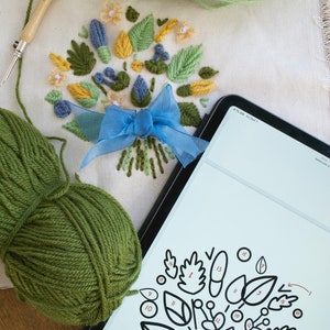 May include: A close-up of a digital tablet displaying a black and white line drawing of a floral design with numbered leaves.  The tablet is lying on a white fabric surface with a green ball of yarn and a partially completed embroidery project featuring a blue ribbon and green, yellow, and blue yarn.