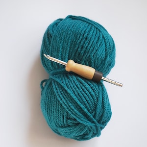 Op de afbeelding: Een bol teal-kleurig garen met een haaknaald erbovenop. De haaknaald heeft een gladde, lichtgekleurde houten handgreep, een donkerbruine kraag en een zilverkleurige metalen schacht met een haak aan het uiteinde en gegraveerde markeringen.