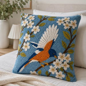 May include: A square throw cushion with a blue background featuring a textured punch needle design of a bird with orange, blue, and white feathers perched on a branch. The design includes white cherry blossom flowers and green leaves.