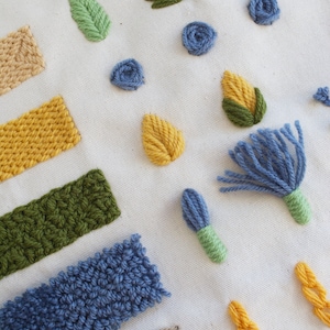 May include: A close-up of a white fabric with various embroidery stitches in different colors, including blue, green, and yellow. The stitches are arranged in a variety of shapes, including leaves, flowers, and geometric patterns.