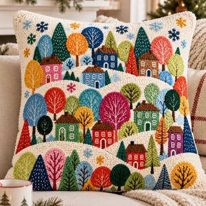 Puede incluir: Un cojín cuadrado con un bordado texturizado estilo punch needle que representa un paisaje invernal. El diseño muestra casas coloridas entre árboles de tonos vibrantes y colinas blancas, decorado con pequeños copos de nieve azules.