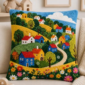 Puede incluir: Un cojín cuadrado con un diseño de paisaje texturizado hecho con aguja mágica. La escena muestra un campo con casas coloridas en rojo, azul y amarillo, colinas verdes, un camino sinuoso, un cielo azul con una nube blanca y un borde floral.