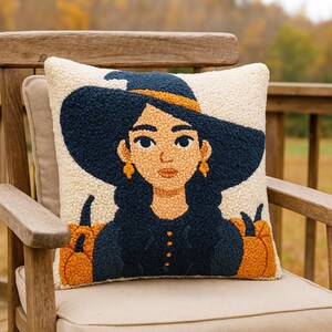 May include: A decorative square pillow featuring a witch illustration. The witch has dark blue hair and a matching hat with an orange band. The pillow has a cream-colored background and two orange pumpkins on each side of the witch.
