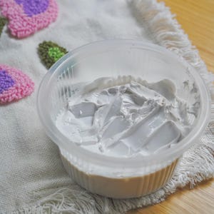 May include: A clear, round plastic container filled with a thick, white, opaque substance that has a textured, swirled surface. The container is placed on a white fabric surface featuring embroidered pink and purple floral patterns.