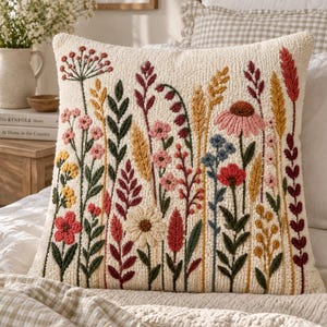Puede incluir: Un cojín cuadrado con base color blanco roto que presenta un diseño bordado en relieve y texturizado de diversas flores silvestres en tonos rojo, rosa, amarillo, azul y verde oscuro, dispuesto en un patrón botánico denso y vertical.
